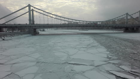 Eine-Gefrorene-Flussoberfläche-Und-Eine-Brücke-Darüber-Inmitten-Einer-Winterlichen-Stadtlandschaft