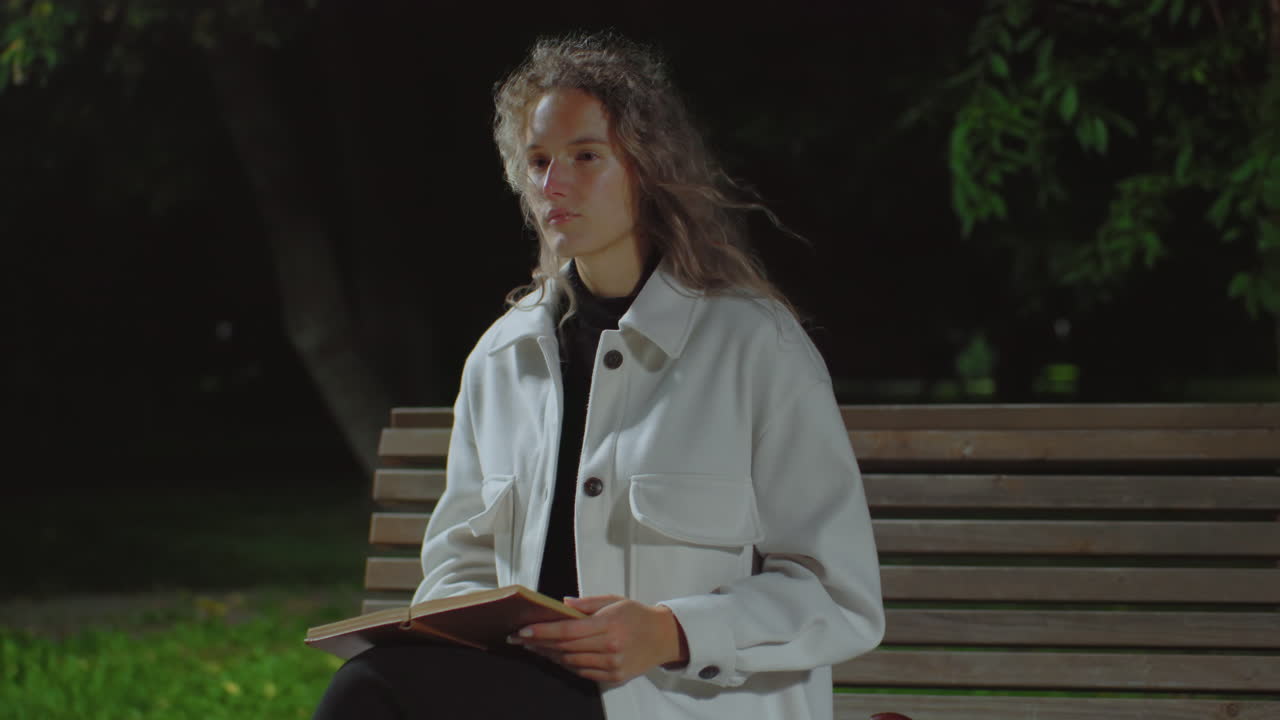 High school student sitting on park bench at night holding open book and reflecting thoughtfully while gentle breeze sways curly hair and nearby tree branch, surrounded by soft dark background