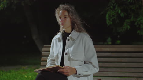 high school student sitting on park bench at night holding open book and reflecting thoughtfully while gentle breeze sways curly hair and nearby tree branch, surrounded by soft dark background
