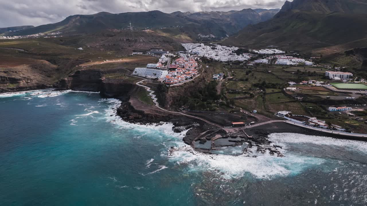 Ocean waves are crashing against the coast of agaete, gran canaria, canary islands, in this beautiful timelapse showcasing the natural swimming pools and the white houses of the town