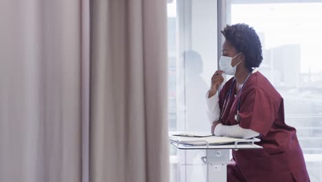 african american female doctor wearing face mask and taking notes in hospital room, slow motion