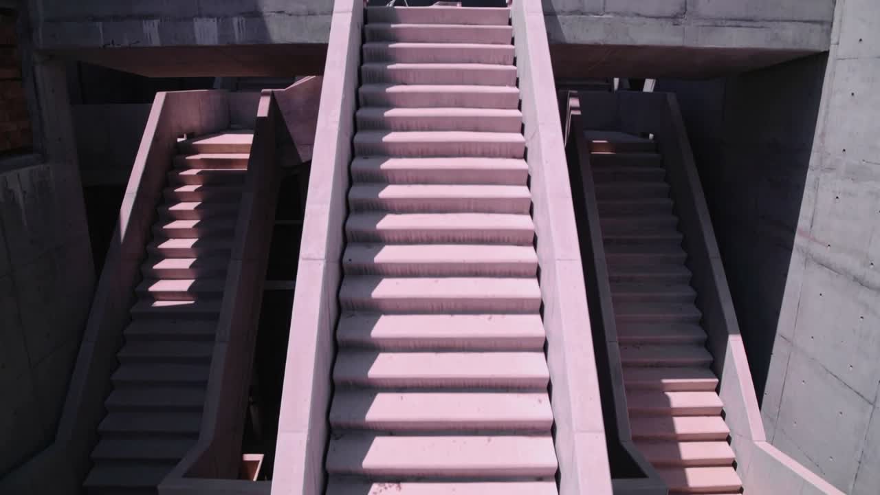 Zoom avant de la caméra débutant sur le palier supérieur, révélant un escalier central nervuré dans une structure en béton avec un faisceau.