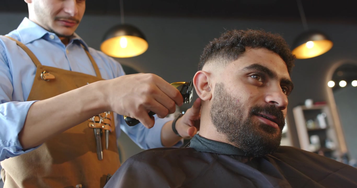 Barbeiro e cliente homem africano inclinados para a direita recebendo corte degradê em barbearia com máquina e pente.