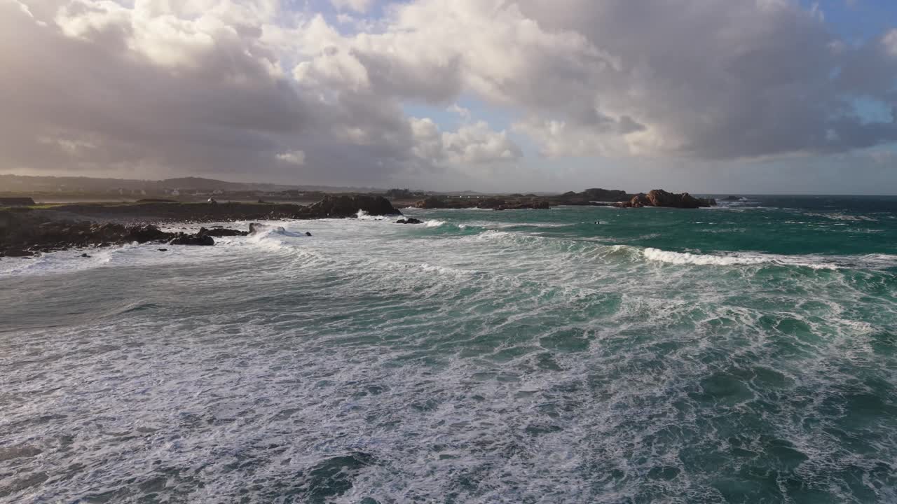 Vuelo lento y bajo al atardecer sobre olas blancas espumosas que rompen en la orilla y se extienden mar adentro, con una luz mágica y un cielo amenazante bajo el sol tardío