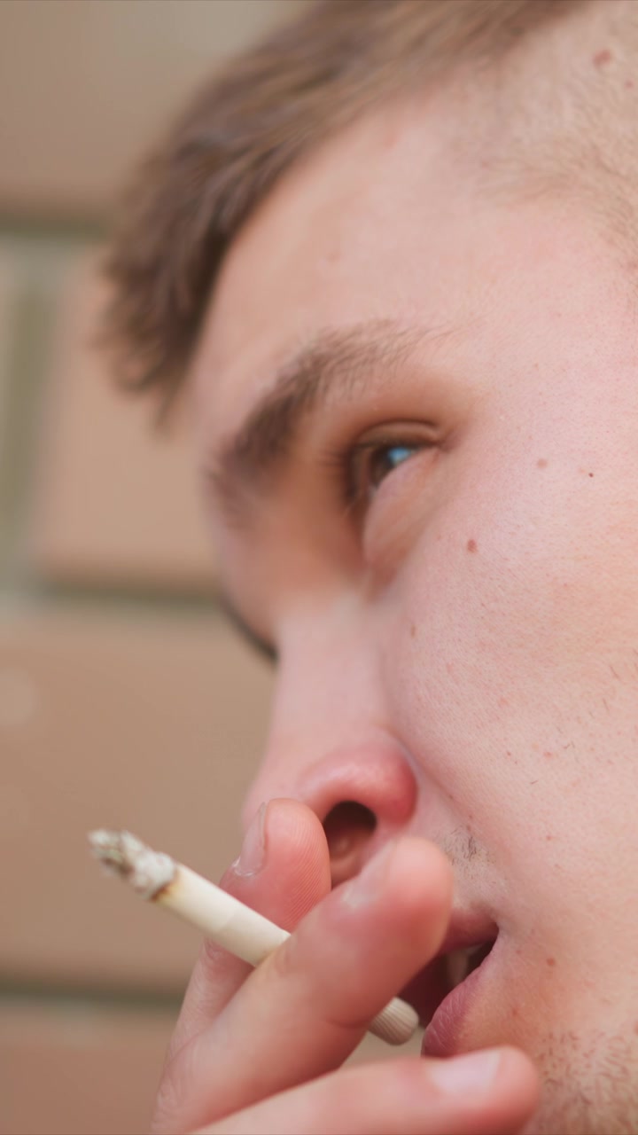 Primer plano de un joven fumando, sosteniendo y llevando un cigarrillo a su boca. La escena se sitúa sobre un fondo de pared de ladrillo desenfocado con una iluminación suave que resalta los rasgos faciales y la piel.