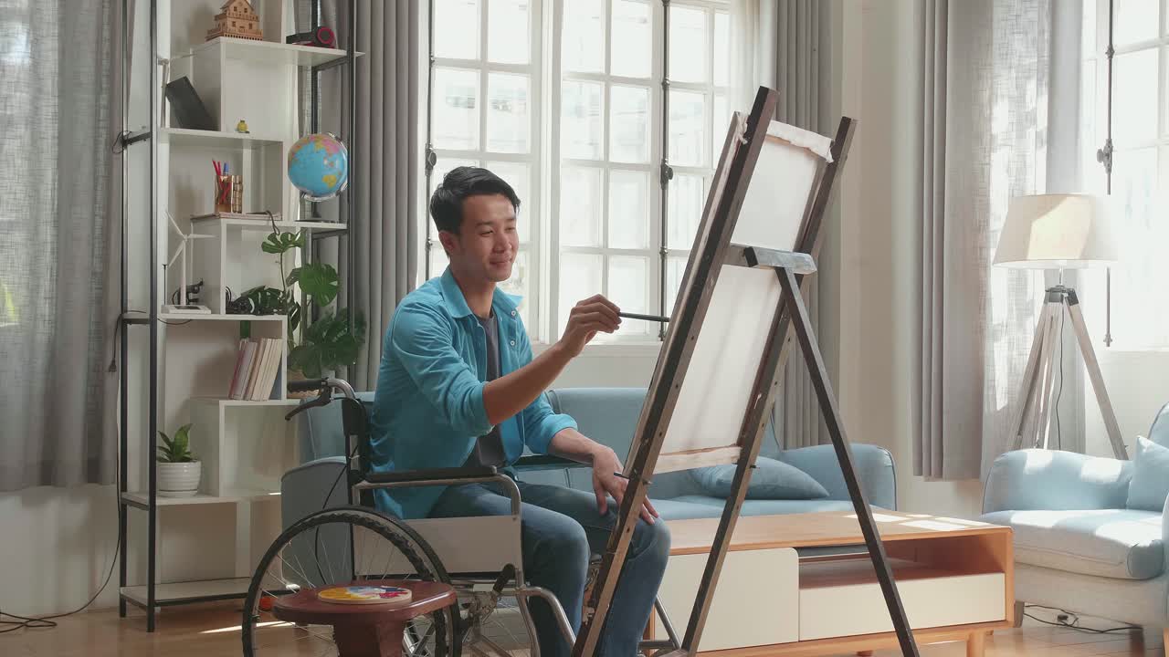 Side View Of An Asian Artist Man In Wheelchair Holding Paintbrush Mixed Colour And Painting On The Canvas