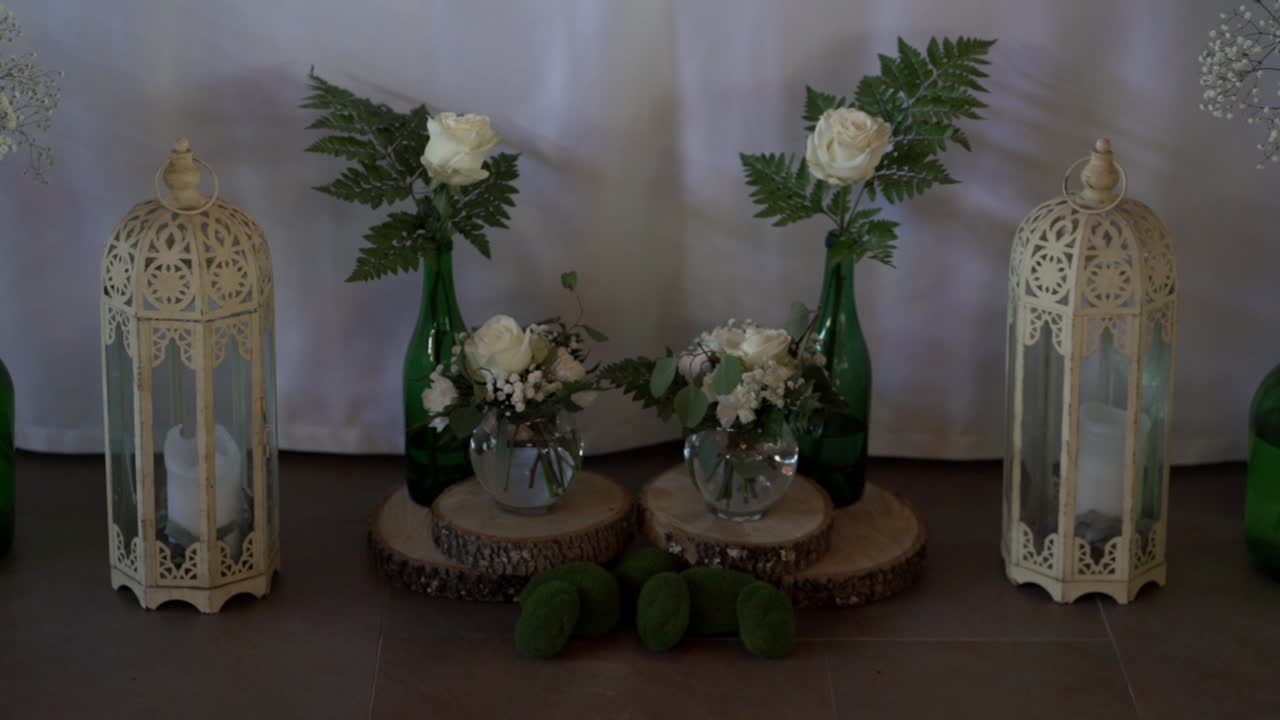 Dekorasi pernikahan pedesaan simetris dengan lilin lentera putih, botol kaca hijau, rangkaian bunga kecil, dan alas irisan kayu di atas meja