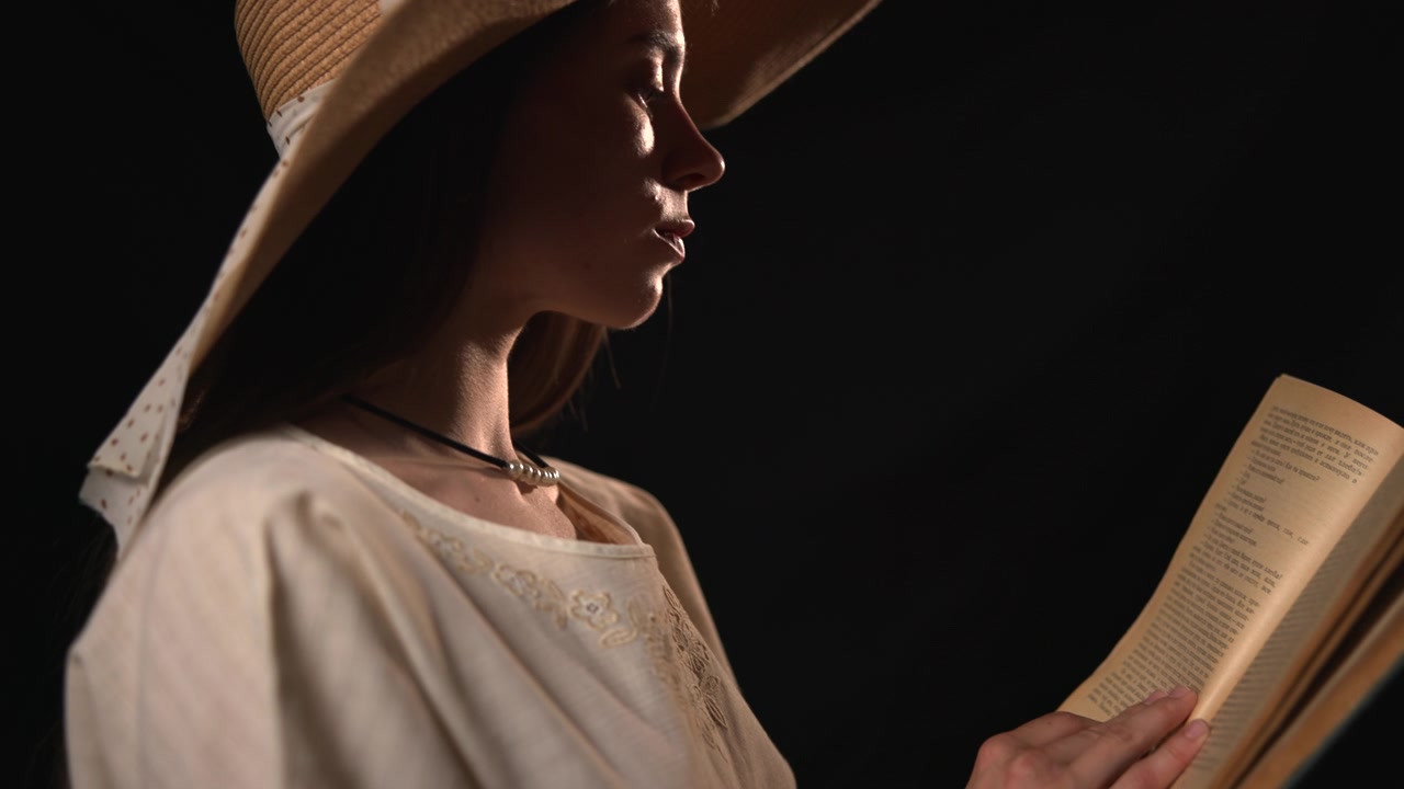 Profilo laterale di una donna adulta che indossa un cappello di paglia e una camicetta chiara, mentre legge un libro aperto in un ambiente interno scuro con un'illuminazione calda e drammatica e un'atmosfera calma.