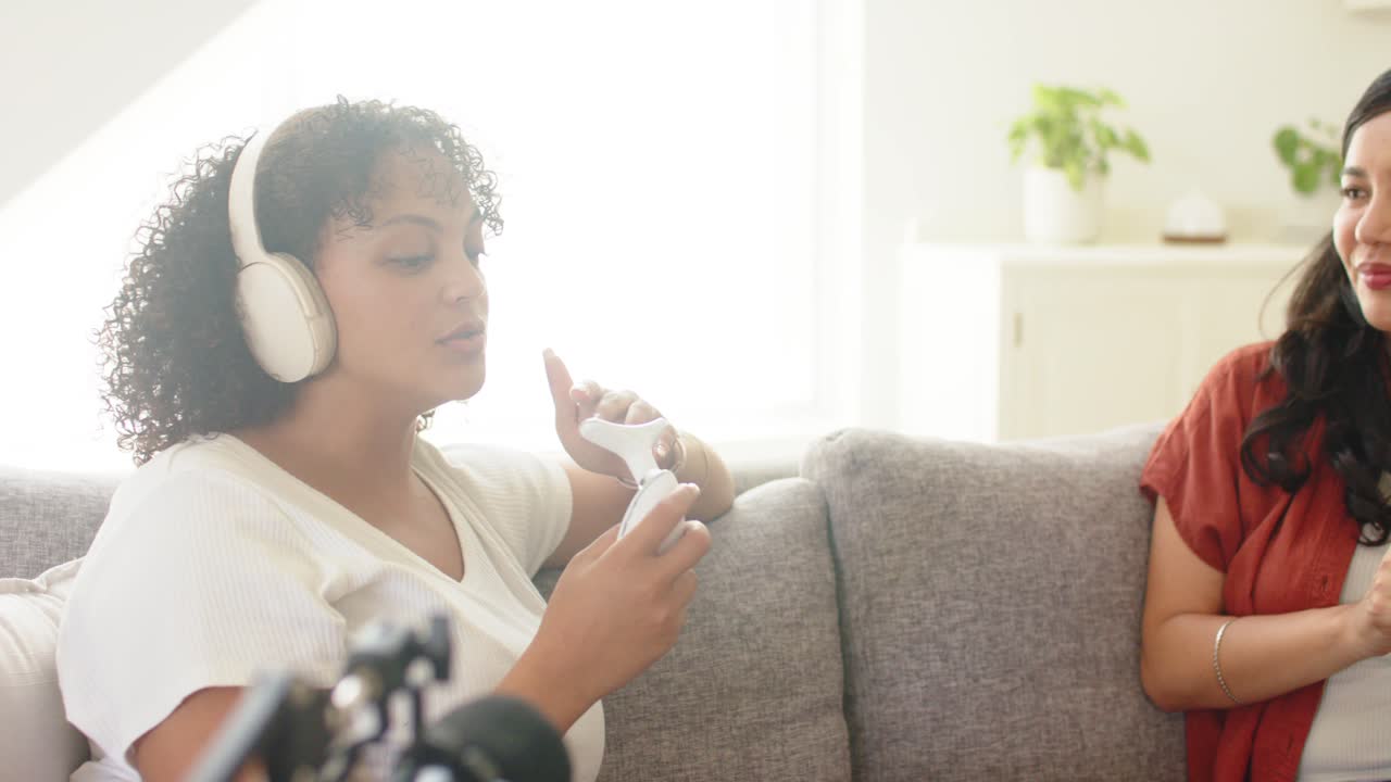 Diverse female cohosts on couch, left showing device, right putting on headphones, recording by mic