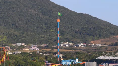 brazilian flag waves atop the vibrant drop tower ride at beto carrero world theme park, with hills and village homes nestled in lush greenery near penha, santa catarina, brazil