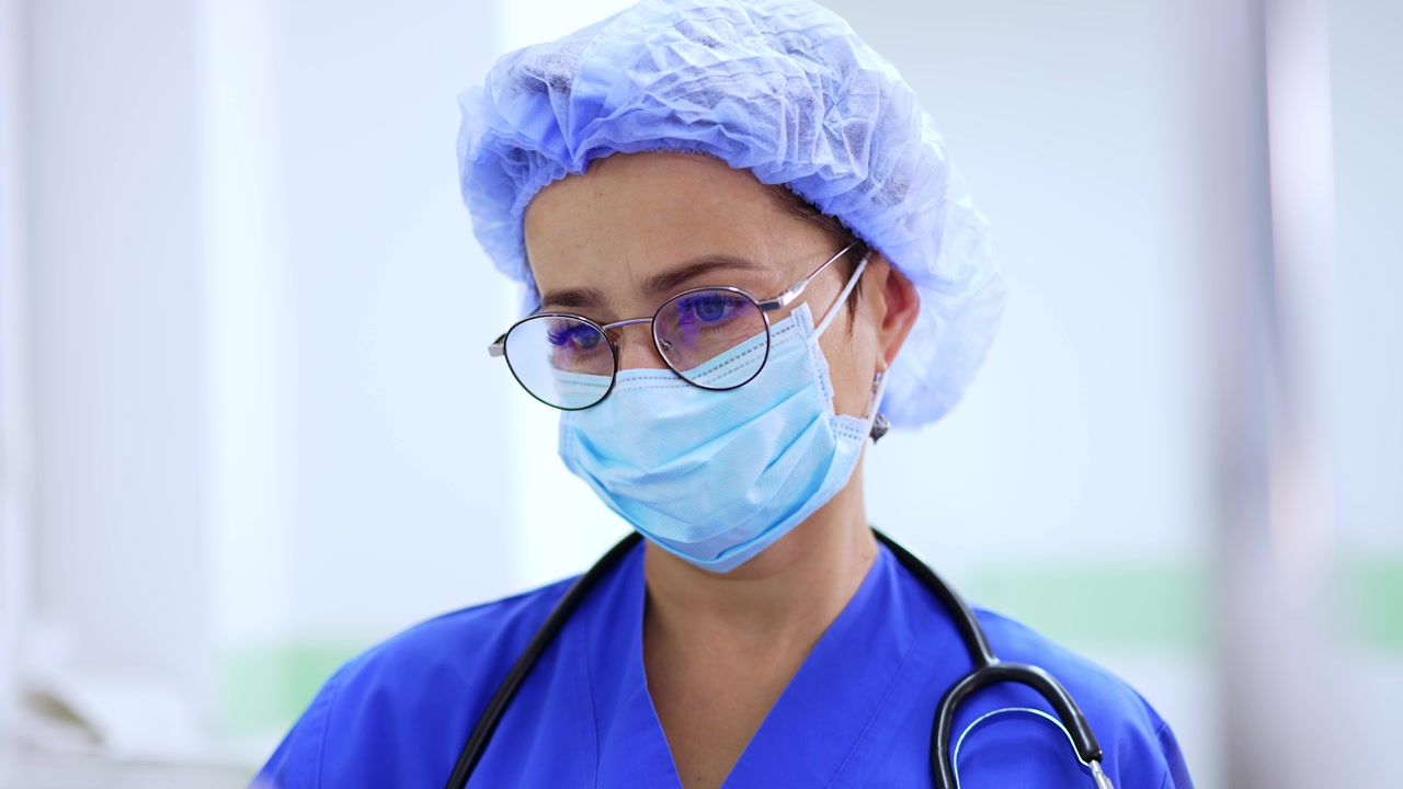 Good-looking Caucasian woman wearing glasses, cap and mask with stethoscope on the shoulders looks intently on the medical apparatus. Medic working in surgery room.