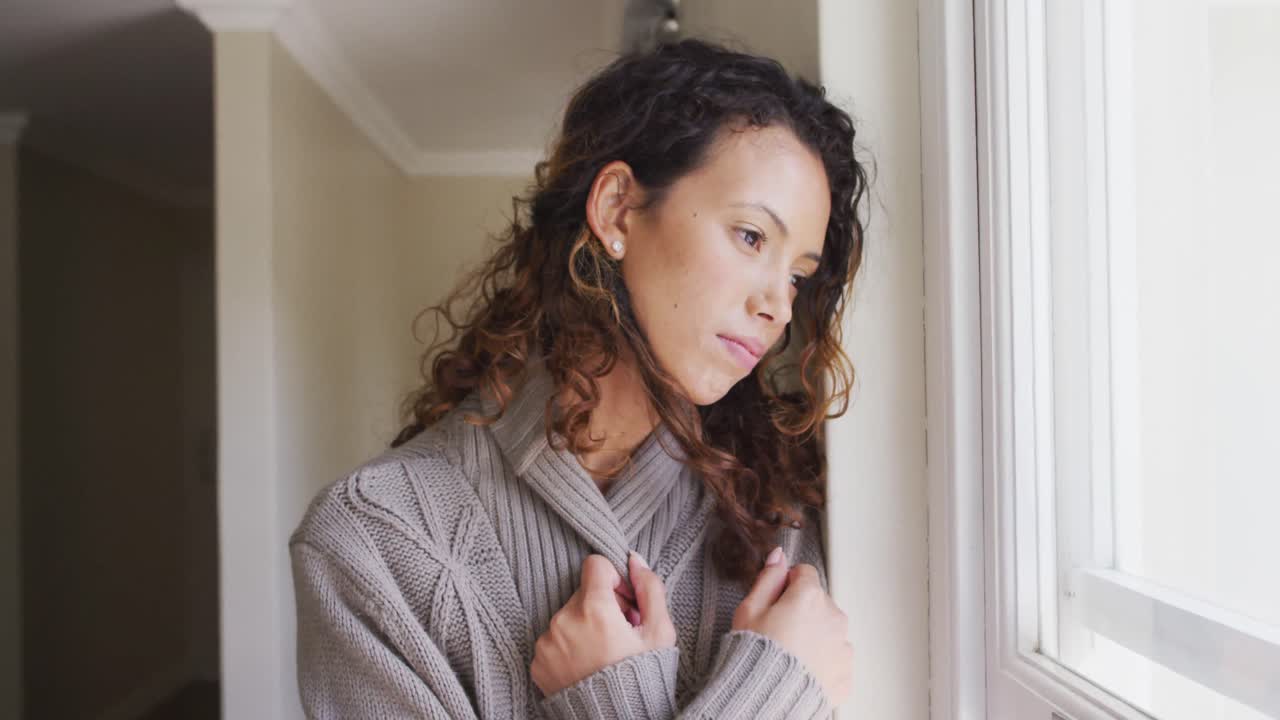 mujer biracial pensativa de pie en la ventana y mirando a la distancia
