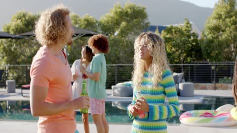 Happy-caucasian-couple-with-drinks-talking-at-sunny-pool-party-with-diverse-friends,-slow-motion