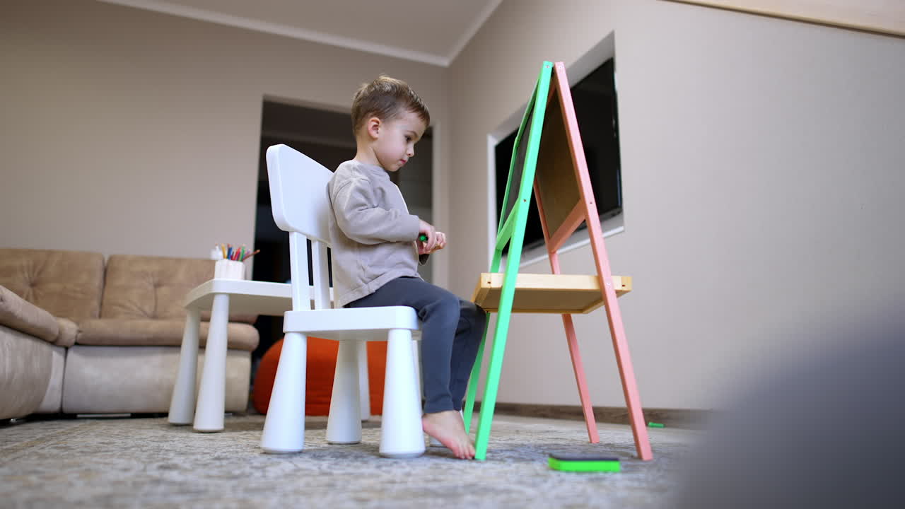 Child draws creatively. A young child sits on a chair focused on drawing at a colorful easel in a cozy indoor space