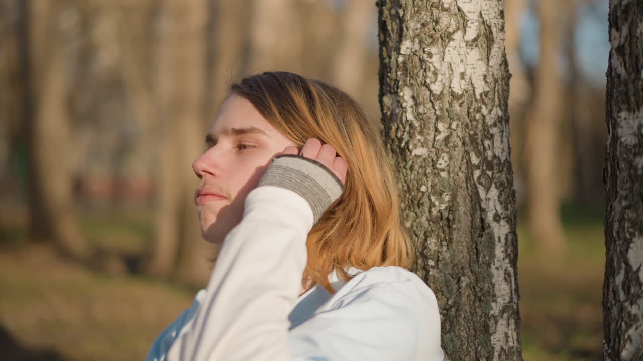 Молодой человек внимательно изучает устройство на запястье рядом с березой, сосредоточенное выражение лица и аккуратные движения рук, залитый солнцем парк на заднем плане и детальные крупные планы подчеркивают техническое любопытство