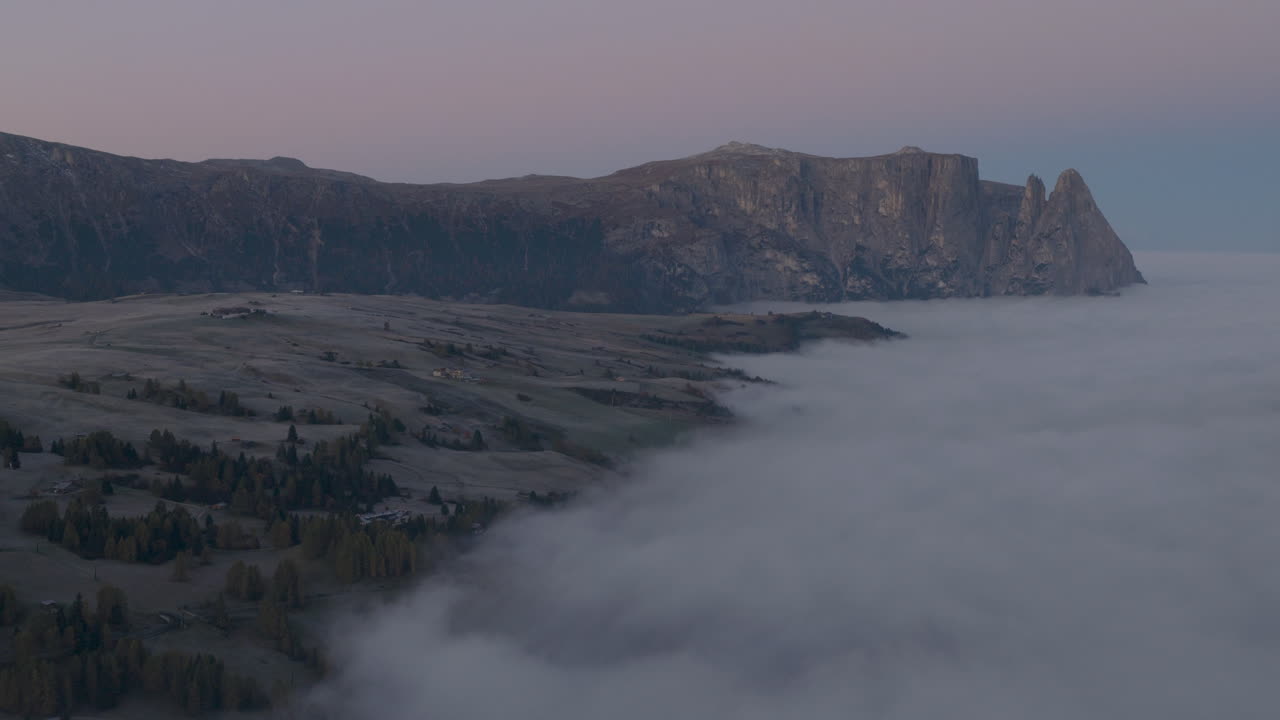 Cinematic aerial view of frosty Alpe di Siusi meadows and scattered mountain huts at dawn, mist drifting across rolling hills beneath the dramatic Sassolungo peaks in the Dolomites, Italian Alps