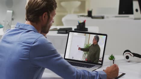 Caucasian-male-student-using-laptop-on-video-call-with-male-teacher,-making-notes