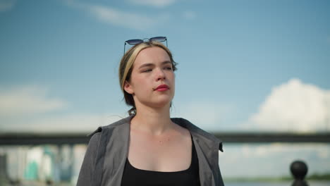 una mujer blanca con una camiseta gris abierta camina pensativa, con un borrón de coches moviéndose en el puente de arriba detrás de ella, bajo un cielo azul claro, parece profundamente pensativa mientras camina