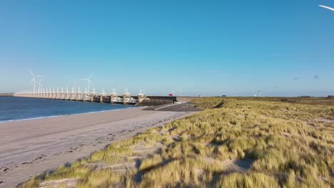 toma aérea sobrevolando dunas, playas y turbinas eólicas hacia la barrera de marejada ciclónica del este de scheldt en zelanda, los países bajos, en un hermoso día soleado