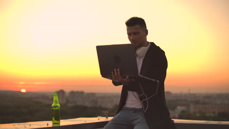 un joven sentado en el borde del techo con una computadora portátil y una cerveza trabajando escribiendo en una computadora portable
