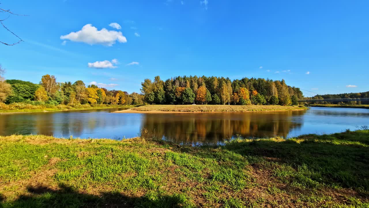 Elv som sakte flyter forbi en skogkledd elvebredd, og viser frem høstfarger under en klar blå himmel i vidvinkel.
