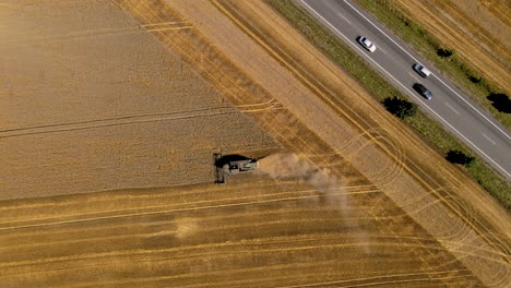 Der-Mähdrescher-Arbeitet-Auf-Dem-Feld,-Mäht-Korn-Nah-An-Der-Straße,-Autos-Fahren-Auf-Der-Straße,-Drehen-Sich-Von-Oben-Nach-Unten-In-Der-Luft