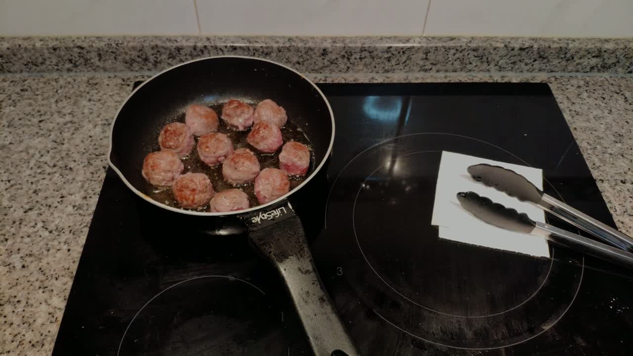 Polpette di manzo e maiale tritato con spezie fritte con olio d'oliva in padella su un piano cottura in ceramica da cucina, con delle pinze accanto, in un primo piano dall'alto che si allontana, Galizia, Spagna.
