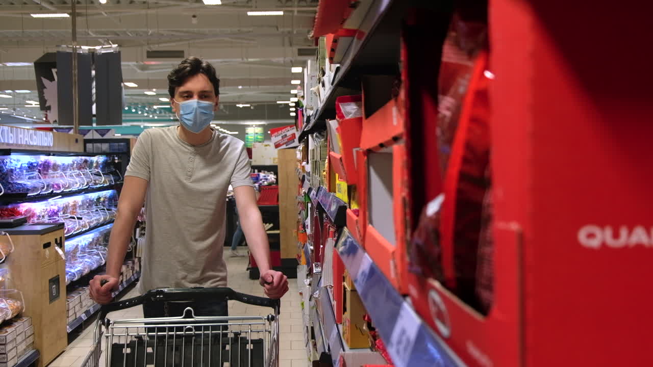 Young man with a protective medical mask walking in a supermarket near the shelves and taking snacks. Corona Virus idea