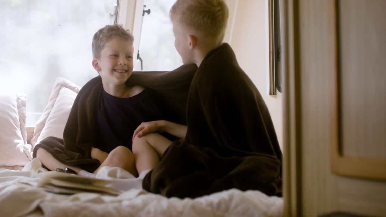 Two little brothers with blanket on their shoulders sitting on bed in a campervan, talking and giving high five