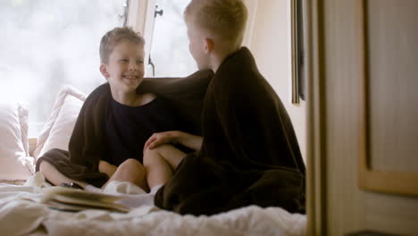 two little brothers with blanket on their shoulders sitting on bed in a campervan, talking and giving high five