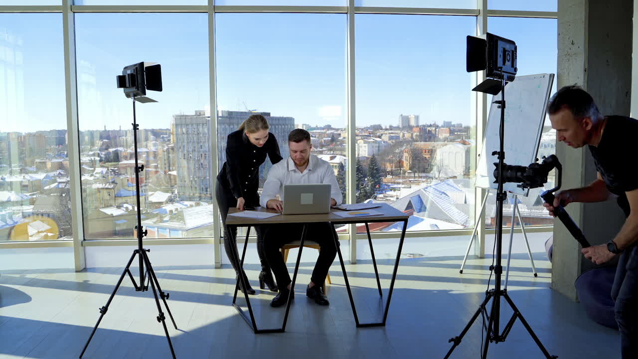 Partners work together in modern office. Businessman works on a laptop and female colleague helping him. Panoramic window view with a city background.