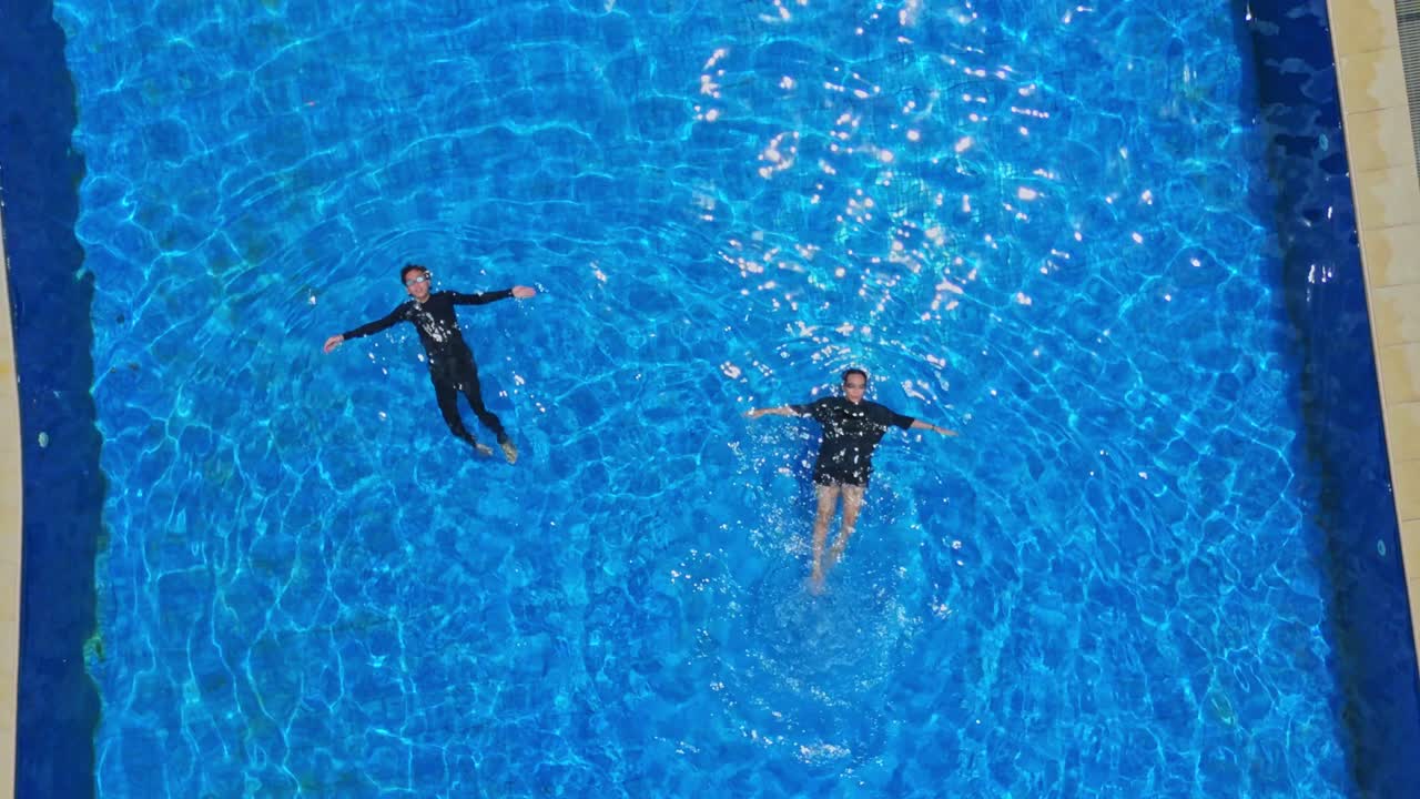 Rekaman drone udara seorang pria dan wanita muda bersantai telentang di kolam renang biru kehijauan yang jernih.