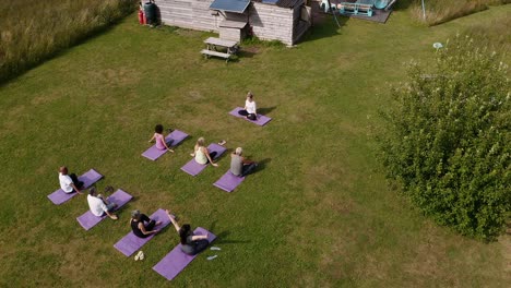 Drone-Shot-Of-Teacher-Leading-Group-Of-Mature-Men-And-Women-In-Class-At-Outdoor-Yoga-Retreat