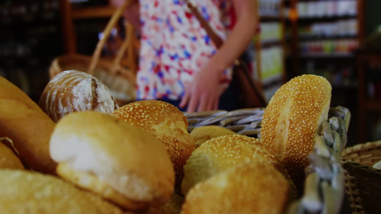 Basket of assorted bread in foreground, shopper hand entering, pressing sesame roll, lifting to buy