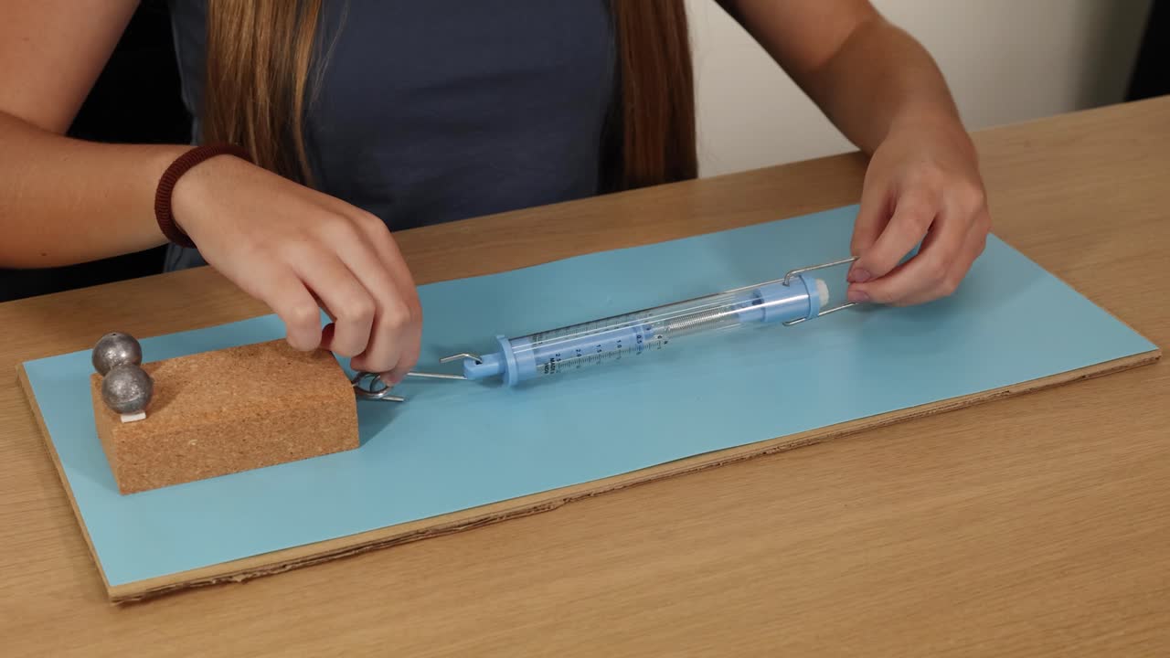 Hands manipulate a spring scale to measure force needed to slide a weighted block on cardboard