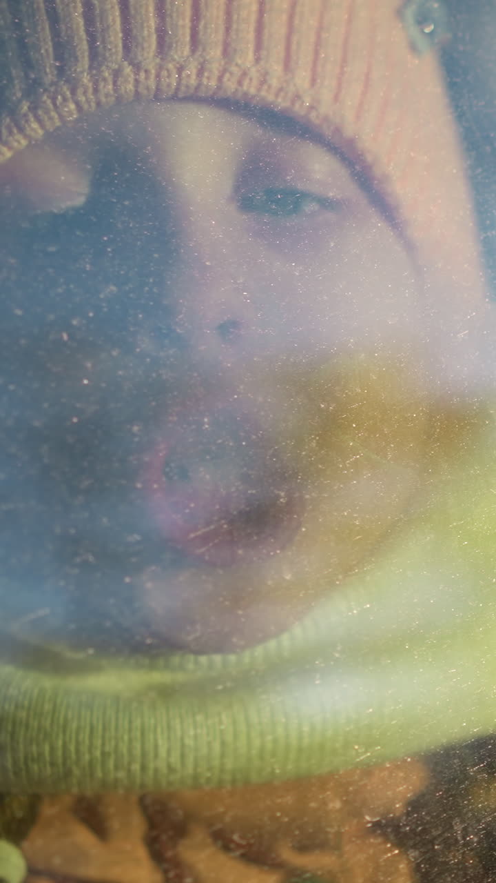 una joven con un sombrero de punto rosado y una bufanda soplando aire en una ventana de coche brumosa, creando condensación. su rostro está presionado contra el vidrio