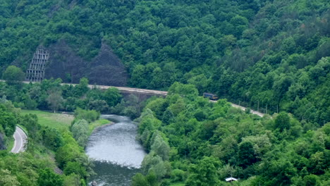 Tracking-wide-shot-of-a-single-locomotive-in-a-beautiful-mountain-passing-near-a-river-in-a-forest