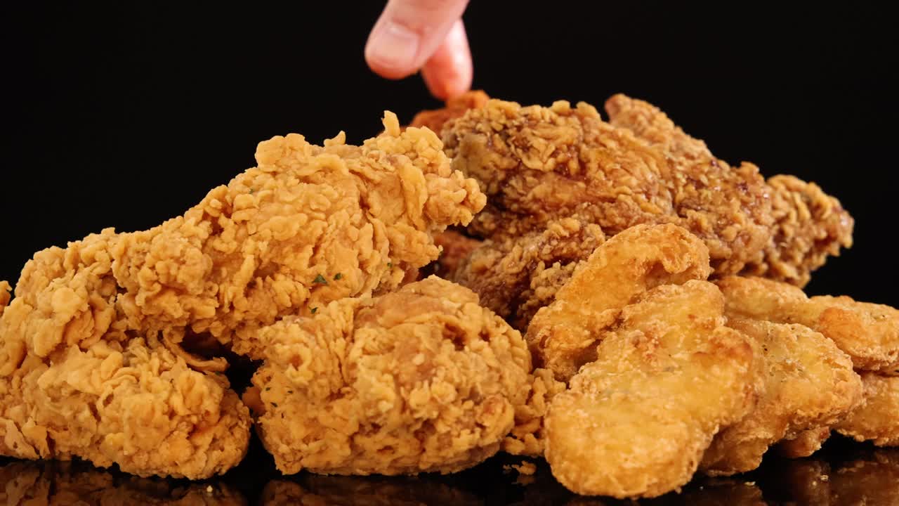 A hand repeatedly picks up pieces from a pile of crispy fried chicken wings, drumsticks, and nuggets on a glossy black surface with dramatic lighting