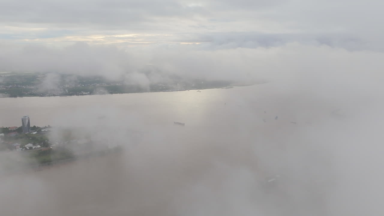 Aerial video of Phnom Penh shows the Tonle Sap river peeking through thick, low-hanging clouds. A few buildings and cargo boats emerge through the fog in a dramatic, cinematic atmosphere