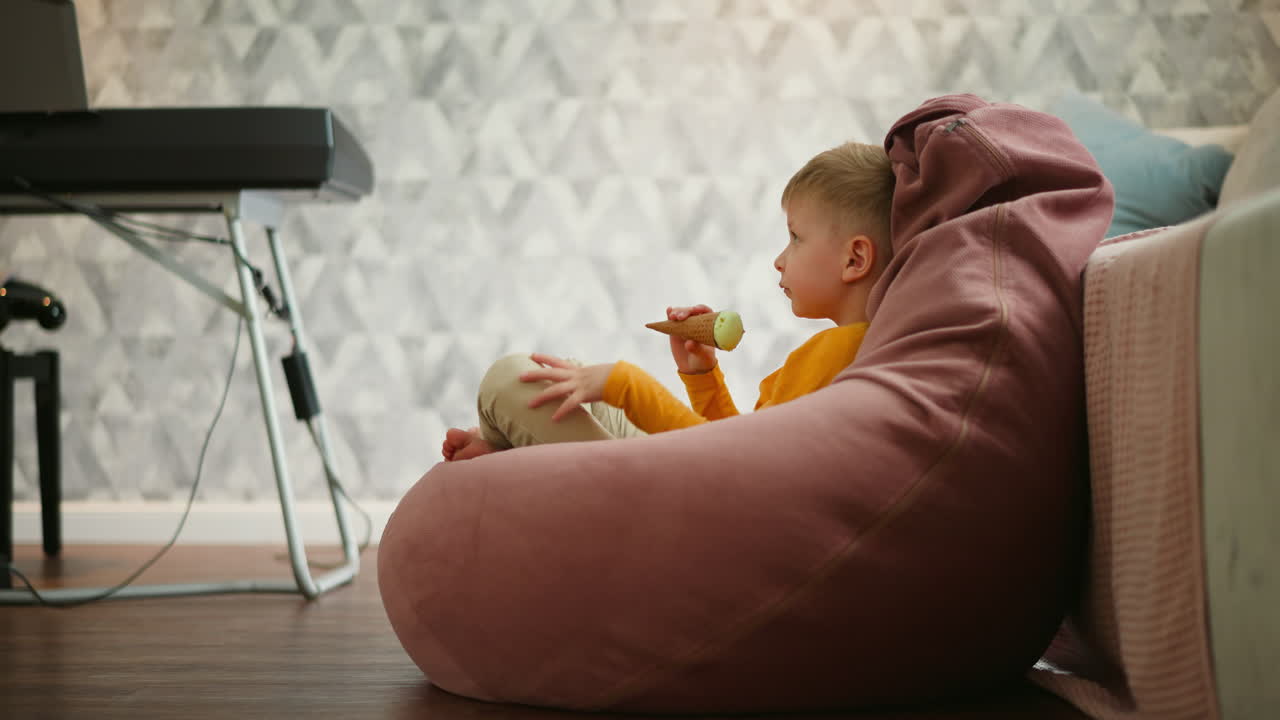 Side view of young boy sitting relaxed on large pink bean bag, holding mint chocolate ice cream cone, gazing ahead with partial view of keyboard and stand in softly lit cozy indoor environment