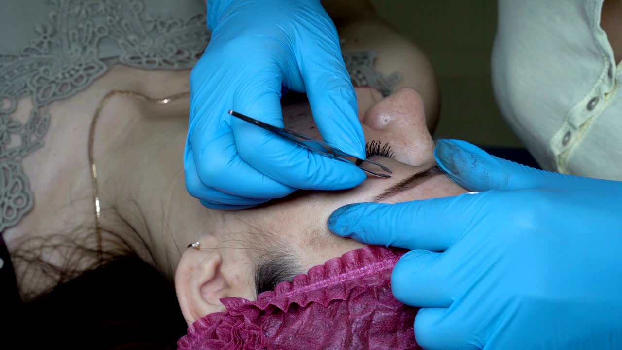 Woman-beautician makes correction of the shape of the eyebrows of the girl in the beauty salon. It plucks and removes small hairs around the eyebrows. Close up. The view from the top. 4K, 25 fps.