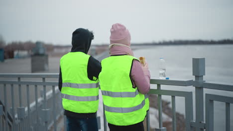 man in hoodie and woman in pink jacket wearing neon safety vests standing side by side leaning on railing overlooking calm river during cold overcast day, peaceful waterfront moment
