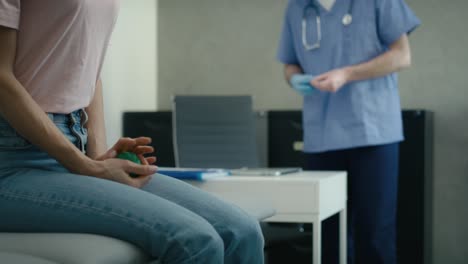 Caucasian-woman-sitting-on-medical-bed-and-squeezing-stress-ball-while-doctor-is-preparing-for-the-medical-exam-in-the-background