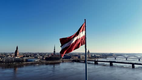 Sunlit-Latvian-flag-in-a-tall-flag-flutters-on-the-edge-of-the-Daugava-river,-in-the-background-the-old-city-of-Riga-and-the-Daugava-bridge-can-be-seen,-in-warm-tones