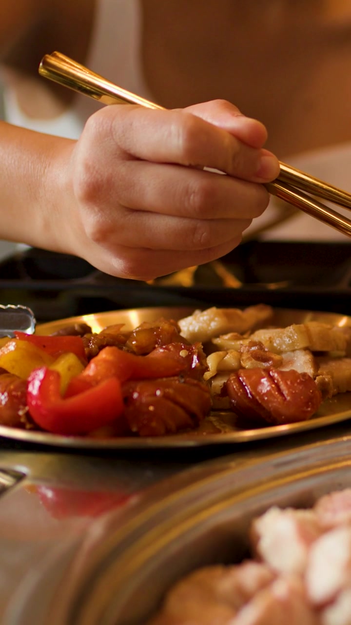 A woman uses golden chopsticks to pick up grilled meat from a brass plate.