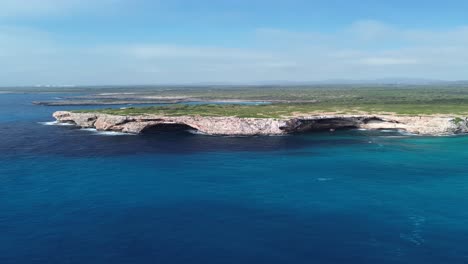 vertical 4k drone footage flying backwards along mallorca’s coastline, revealing two large sea caves, rocky cliffs, green bushes, and crystal clear blue water. captured on a sunny day in june 2025