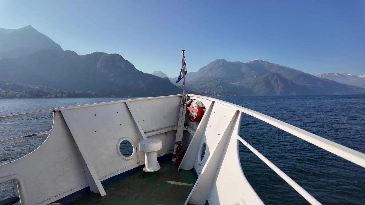 Vista dal ponte di prua da un traghetto che attraversa il Lago di Como in Italia, con acqua di un blu intenso, paesaggi montani spettacolari e cieli sereni all'orizzonte