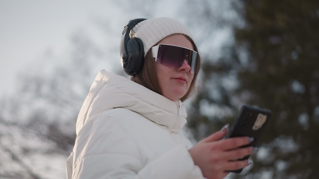 Happy young woman pressing mobile phone with smile on face while wearing tinted goggles headphones and winter coat in snowy park under soft sunlight capturing joyful moment and blurred tree backdrop