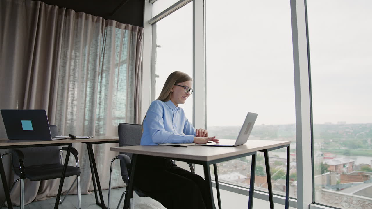 Busy lady having online chat on her laptop. Smiling woman working on her computer near the big panoramic window with cityscape.