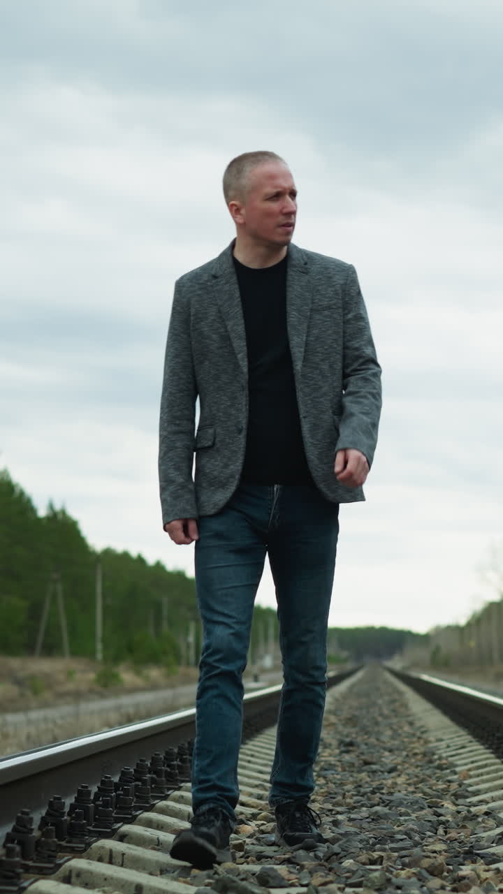 A close view of an aged man wearing jeans, a grey suit, and canvas shoes, walking tiredly on a railway with stones, with trees and electric poles on either side